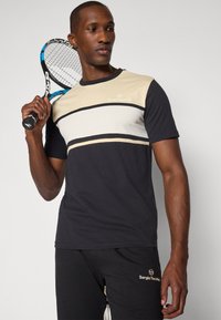 Camiseta negra de hombre de manga corta con rayas horizontales en crema y beige claro y logo en el pecho, combinada con pantalones negros.