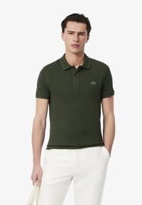 Polo shirt verde scuro in cotone, con patta a due bottoni, maniche corte e un piccolo logo ricamato sul petto. Indossato con pantaloni bianchi.