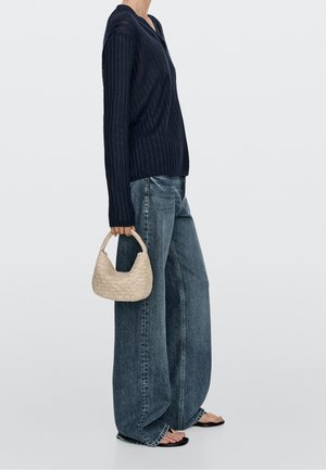 Frau trägt schwarzen gerippten Pullover und Jeans mit weitem Bein, hält eine kleine gewebte Handtasche und steht vor weißem Hintergrund.