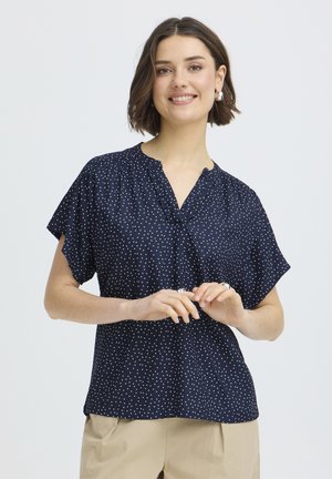 Usmievajúca sa žena s krátkymi hnedými vlasmi, ktorá má na sebe strieborné kruhové náušnice, tmavomodrú blúzku s krátkym rukávom a biele bodky a béžové nohavice.