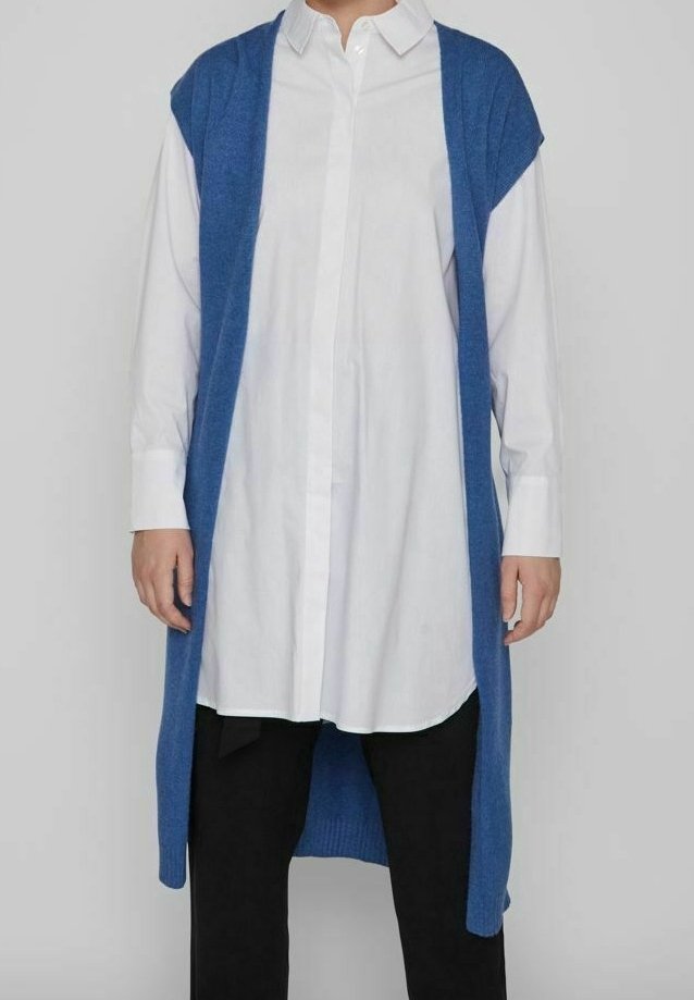 Une personne porte un long cardigan bleu sans manches par-dessus une chemise blanche boutonnée et un pantalon noir, debout devant un fond clair uni.
