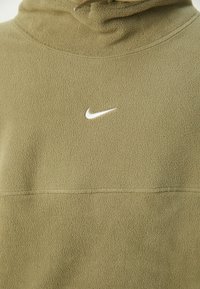 Gros plan d'un pull polaire vert olive avec le logo Nike Swoosh blanc sur la poitrine et un col montant avec un cordon.