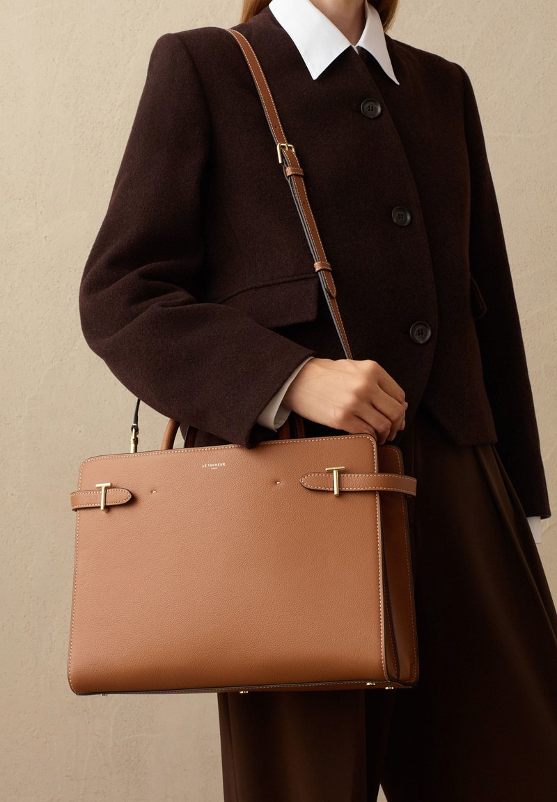 Bolso de mano de cuero marrón con herrajes dorados, asas dobles y correa de hombro ajustable. Presenta un diseño minimalista y un acabado texturizado.