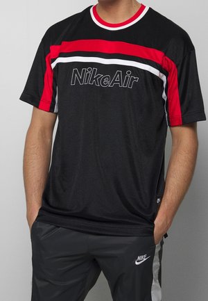 Persoon draagt een zwart NikeAir-shirt met rode en witte strepen, zwarte Nike-broek met wit logo, handen in zakken, staand tegen een grijze achtergrond.