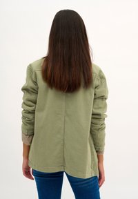 Person with long straight dark hair wearing an olive green jacket and blue jeans, standing with their back to the camera.