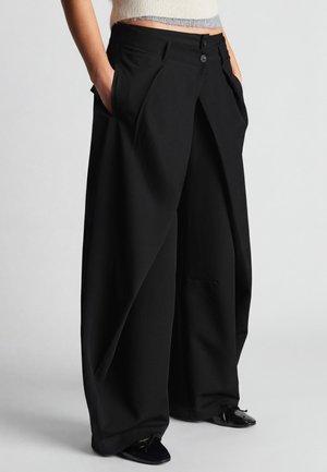 Vrouw draagt zwarte, wijde broek met hoge taille, voorplooien en zakken, gecombineerd met zwarte puntige schoenen en een korte trui.