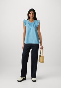 Top azzurro chiaro con maniche a volant, vestibilità ampia, abbinato a pantaloni scuri e sartoriali e una piccola borsa gialla con superficie texturizzata.