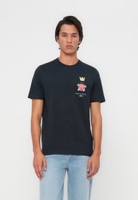 Navyblauwe katoenen t-shirt met ronde halslijn, met een grafisch ontwerp van een kroon en roos op de linkerkant van de borst. Het model draagt lichtblauwe jeans.
