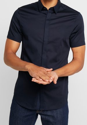 Mann trägt ein eng anliegendes, kurzärmeliges, marineblaues Hemd mit Kragen und Knopfleiste, kombiniert mit dunkelblauen Jeans, die Hände ineinander verschränkt.