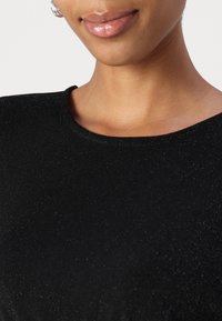 Haut noir texturé avec un col rond et un effet de brillance subtil, révélant une peau lisse sur le cou et les lèvres.