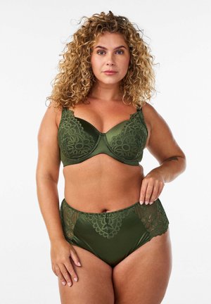 Femme aux cheveux bouclés portant un ensemble de lingerie en dentelle verte, debout devant un fond blanc, regardant la caméra avec confiance.