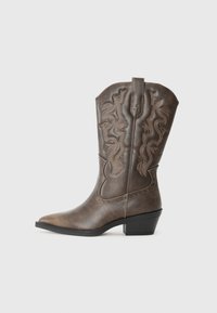 Botas vaqueras de cuero marrón con puntera puntiaguda, costuras decorativas y acentos con tachuelas. Tacón bajo y caña alta para un estilo elegante.