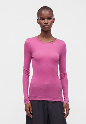 Femme aux cheveux courts portant un t-shirt rose ajusté à manches longues et un pantalon noir ample, debout devant un fond clair uni.
