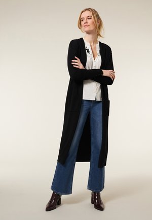 Zwarte lange cardigan met open voorkant, zijzakken, gecombineerd met een witte textuurshirt, blauwe wijde jeans en bordeauxglanzende leren laarzen.