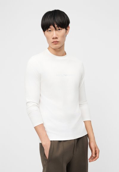 Νεαρός άνδρας με μαύρα μαλλιά που φοράει εφαρμοστή λευκή μακρυμάνικη μπλούζα και καφέ παντελόνι, όρθιος με το ένα χέρι στην τσέπη, μπροστά σε απλό φόντο.