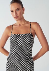 Žena s hladko zpečenými vlasmi, ktorá nosí zlaté kruhové náušnice a čiernobiele šaty bez rukávov s geometrickým vzorom, stojí so založenými rukami za chrbtom.