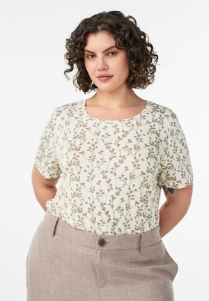 Kvinde med krøllet hår iført en hvid kortærmet skjorte med et lille brunt blomsterprint og beige højttaljede bukser.