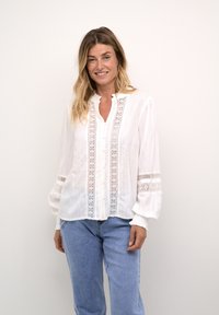 Vit skjorta med korta ärmar och spetsdetaljer fram och på ärmarna. Utsvängda ärmar med elastik vid ärmsluten. Bärs med ljusblå jeans.
