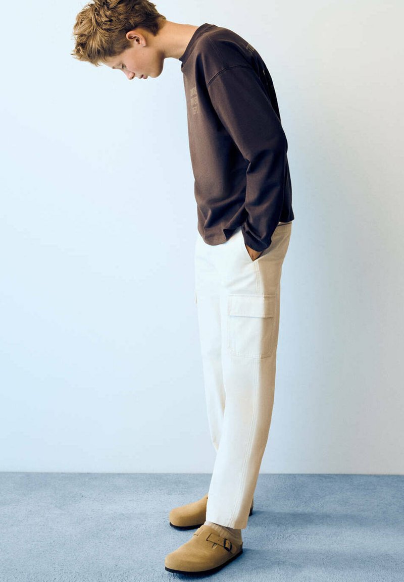 Personne aux cheveux courts portant une chemise à manches longues foncée, un pantalon cargo blanc et des chaussures beige, se penchant en avant les mains dans les poches.