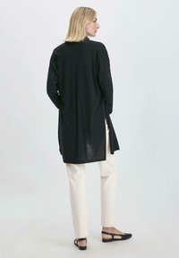 Pitkä musta tunika, jossa on rento istuvuus ja sivuhalkiot, yhdistettynä vaaleanbeigeihin housuihin ja mustiin litteisiin sandaaleihin, yksinkertaisella muotoilulla ja rakenteella.