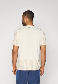 Vīriešu īsām piedurknēm krekls gaiši bēšā krāsā ar horizontālām svītrām sānos un vienkrāsainu aizmuguri. Tam ir apaļa apkakle un smalks logotipa detaļas.