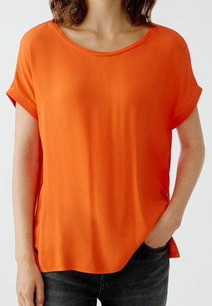 Femme portant un chemisier orange vif à manches courtes avec un col rond, associé à un jean foncé, main dans la poche sur un fond uni.