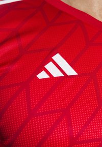 Gros plan sur un tissu de vêtements de sport rouge avec un motif géométrique et un logo Adidas blanc sur la poitrine.