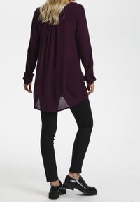 Långärmad, djup lila tunika med lös passform, texturerad tyg och sidoklipp. Bärs med svarta slim-fit byxor och kraftiga svarta skor.