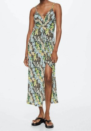 Femme portant une robe maxi sans manches à motifs floraux avec une fente haute à la cuisse et des sandales compensées noires, debout devant un fond blanc uni.