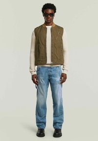 Gilet verde in tela con tasche laterali, colletto beige, indossato sopra un maglione di maglia crema e jeans blu strappati, completato da stivali neri.