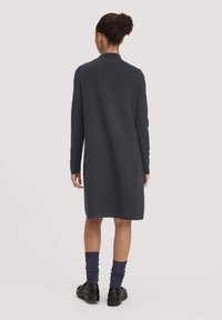 Dunkelgraues, langärmliges Kleid mit geradem Schnitt, aus weichem Stoff. Getragen mit dunkelblauen gerippten Socken und schwarzem Schuhwerk.