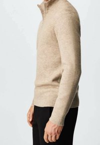 Uomo che indossa un maglione a mezze zip beige a coste e pantaloni neri, in piedi di lato su uno sfondo chiaro e uniforme.
