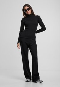 Maglia nera a maniche lunghe con colletto a cratere abbinata a pantaloni neri a campana. Il tessuto sembra morbido con una texture liscia. Indossata con scarpe da ginnastica grigie.