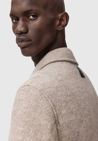 Collaret beige uld frakke med en struktureret overflade og sorte læderdetaljer på ryggen. Model kigger over skulderen og viser beklædningen frem.