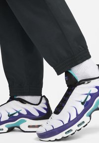 Las zapatillas Nike Air Max presentan un diseño en blanco y morado, con una parte superior de malla, cordones negros y detalles en verde azulado. Se pueden ver pantalones negros con puños elásticos.