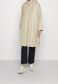 Parka beige con cappuccio, chiusura zip frontale e grandi tasche. Maniche lunghe con polsini. Indossato sopra pantaloni neri e sneakers neutre. Texture liscia.