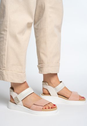 Füße in beige-rosa offenen Sandalen mit weißen Riemen und Sohlen, kombiniert mit hellbeigen, hochgekrempelten Cordhosen.