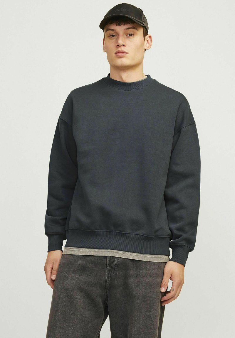 Tumesinine sweatshirt ümmarguse kaeluse ja soonikkoesvarrukatega, mille peal on helesinine särk. Näha on paar tumedaid teksaseid.
