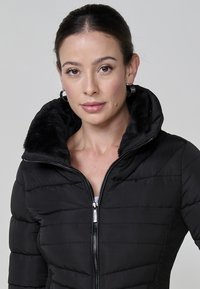 Chaqueta acolchada negra con cremallera frontal, que cuenta con una capucha forrada de pelo sintético y textura acolchada. Paneles laterales en un material de contraste.