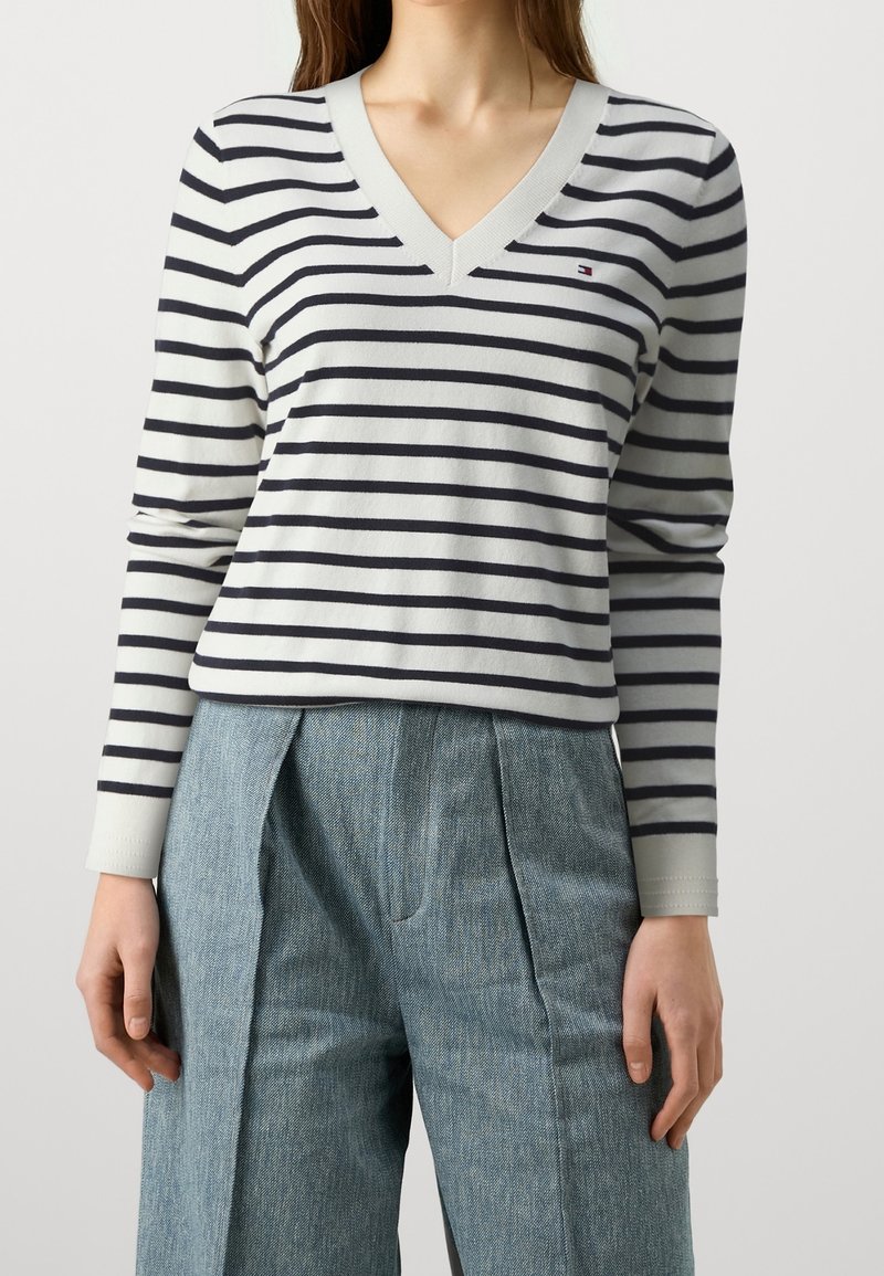 Femme portant un pull à col en V à rayures horizontales blanches et bleu marine et un pantalon ample bleu-gris, debout devant un fond uni.