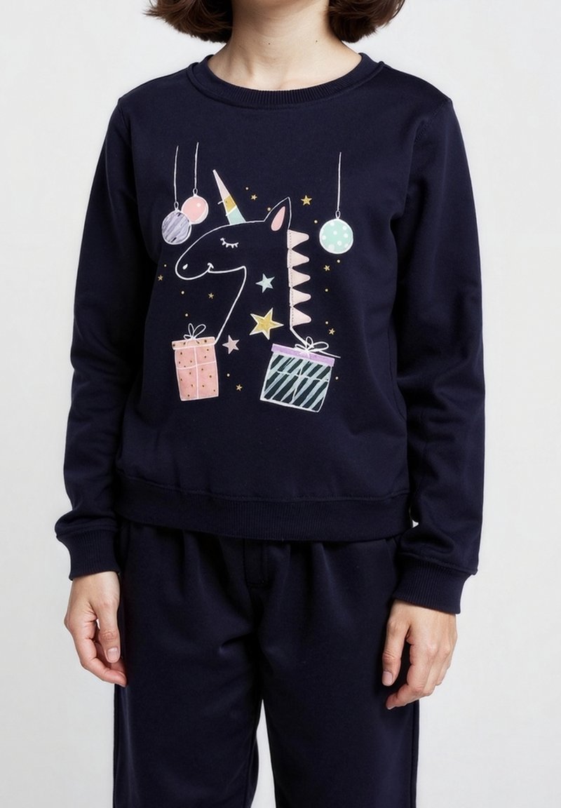 Personne portant un sweat-shirt bleu marine avec des illustrations de licorne, d'étoiles, de décorations suspendues et de deux cadeaux emballés sur le devant.