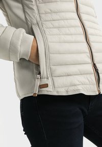 Lysebeige dunjakke med quiltet design, sidelommer med lynlås, ribstrikkede manchetter og en brun læderlogo-patch. Slank pasform med en glat tekstur.