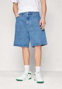 Denim shorts in light blue, featuring a relaxed fit, two side pockets, and a button closure. Paired with white sneakers and socks.