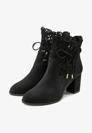 Schwarze Ankle-Boots aus Wildleder mit einem Schnür-Ausschnittdesign, akzentuiert durch eine Schleife und goldfarbene Hardware. Blockabsatz für Stabilität.