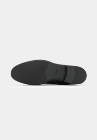 Semelle de chaussure habillée noire avec surface texturée antidérapante et petit logo embossé "BOSS" près de la zone de la voûte plantaire, présentée sur un fond blanc uni.