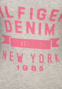 Grå tyg t-shirt med djärv rosa text som säger "ALFIEGA DENIM NEW YORK 1985" med en ribbaccent under. Mjuk struktur.
