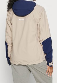 Chaqueta impermeable beige con capucha y mangas en color marino, que cuenta con puños ajustables y el nombre de la marca "Mammut" impreso en la parte trasera.