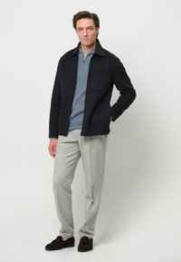 Giacca di lana blu navy con colletto e taschino sul petto, indossata sopra un maglione a coste blu chiaro, abbinata a pantaloni grigi chiari e scarpe scure.