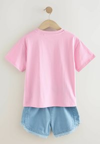 Roze katoenen T-shirt met korte mouwen, gecombineerd met lichtblauwe geribde shorts met een golvende kantafwerking, hangend aan een houten hanger.