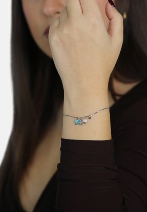 Delicato braccialetto d'argento con turchese, perle e piccoli ciondoli indossato al polso, mano che copre parzialmente il volto, capelli lunghi e scuri visibili.